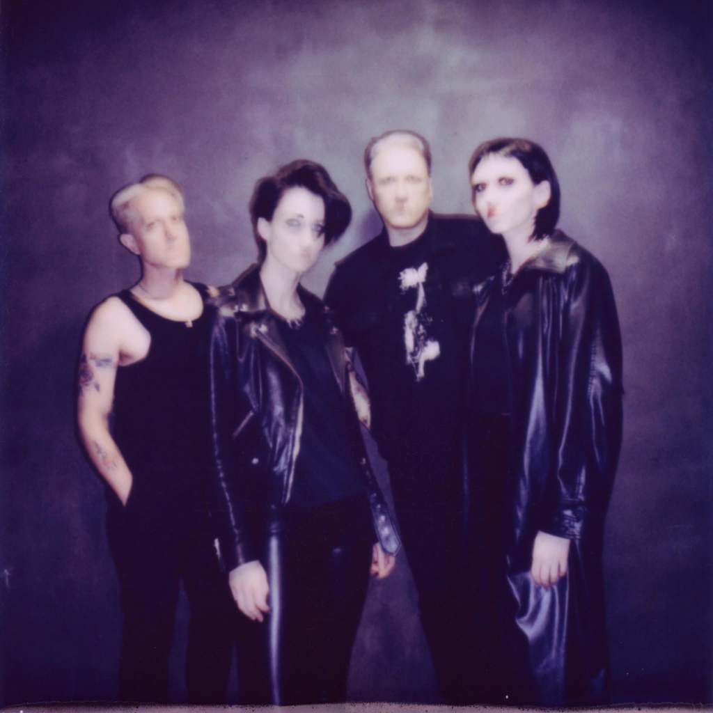 A group of four individuals posing together, dressed in dark, edgy fashion, against a muted background.