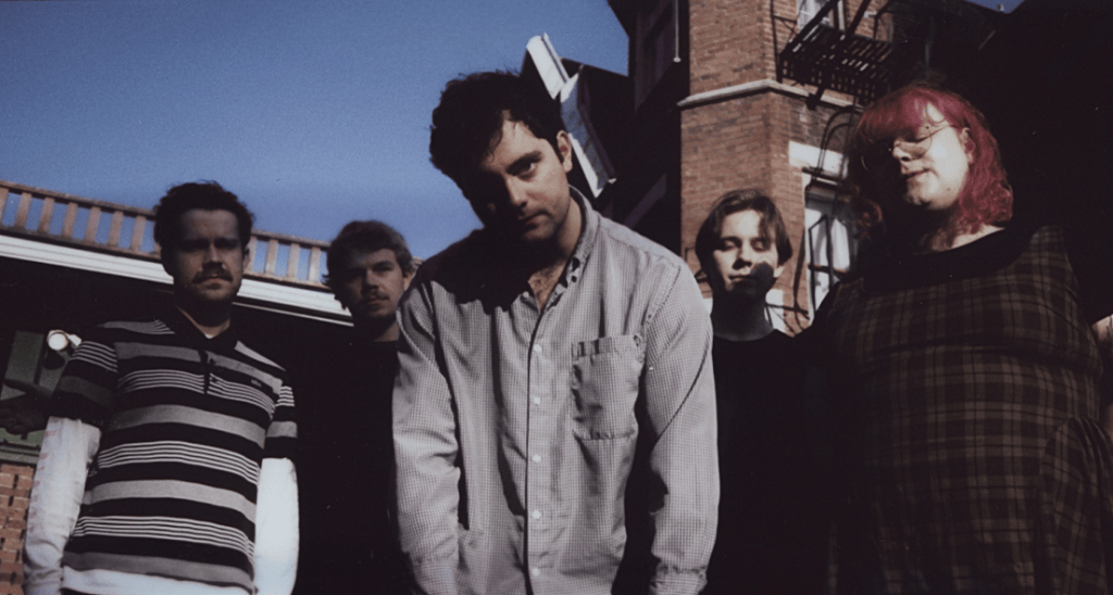 A group of five people posing outdoors, standing close together with a brick building and a blue sky in the background. They have a mix of casual clothing styles and expressions.