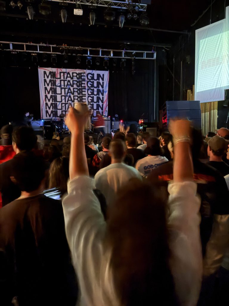 Crowd at a concert with hands raised, enjoying a live performance, featuring a backdrop with 'MILITARY GUN' text.