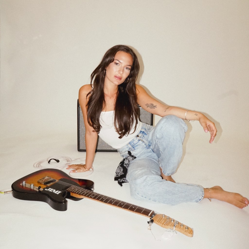 A woman sitting on the floor beside a guitar, wearing a white tank top and light blue jeans, with an amplifier in the background.