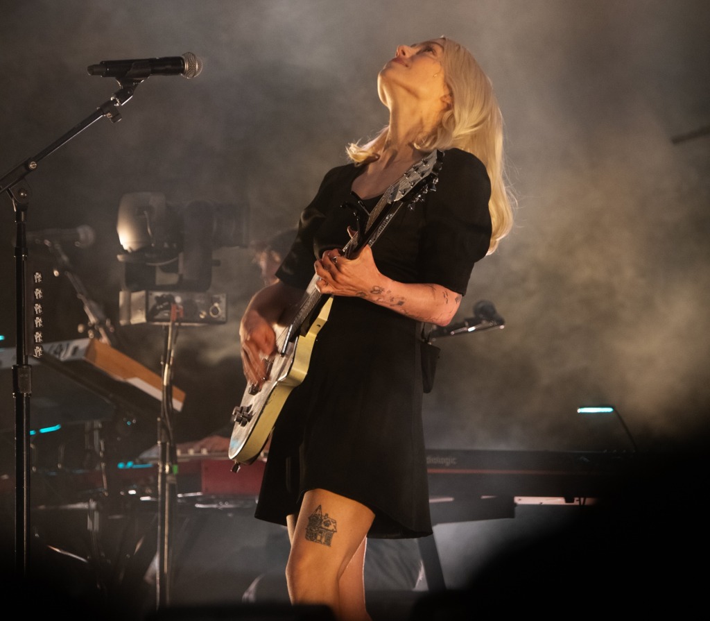 A female musician playing an electric guitar on stage, dressed in a black dress, with long blonde hair and visible tattoos, surrounded by atmospheric lighting and smoke.