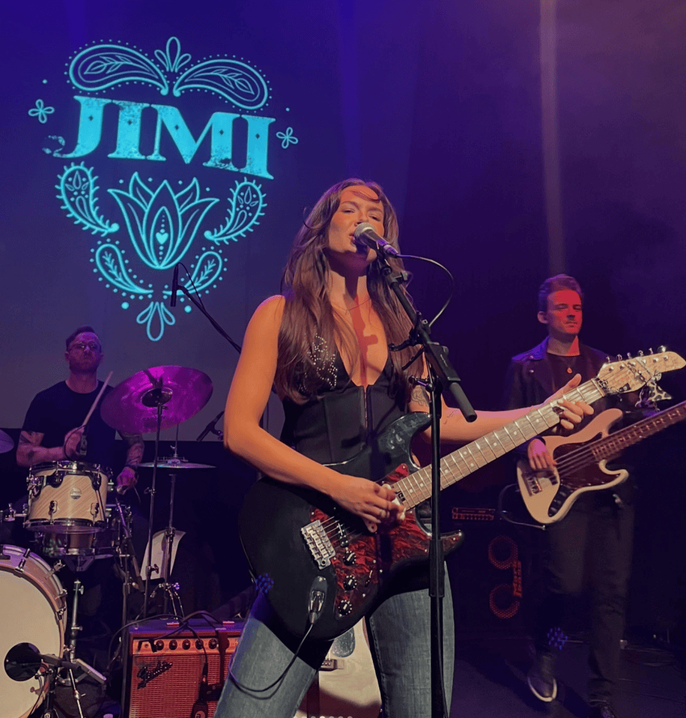 A female musician performing on stage with an electric guitar, singing into a microphone, while a drummer is seen in the background alongside a bassist.