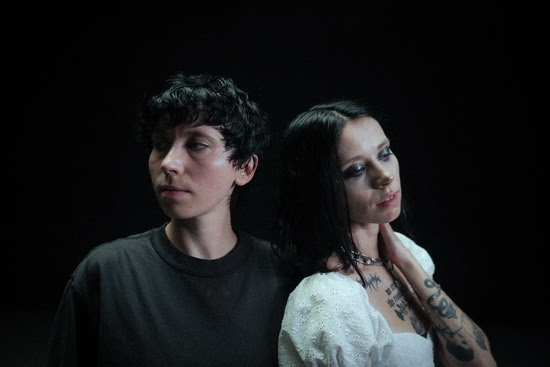 Two individuals standing back to back in a dimly lit space. One has short, curly hair and is wearing a black shirt, while the other has long, dark hair in a white dress decorated with puff sleeves.