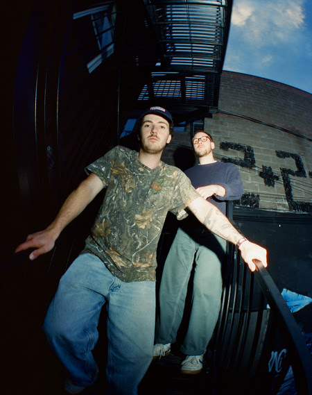 Two young men descending a staircase outside a building, one wearing a camouflage t-shirt and the other in a navy sweater, with a graffiti wall in the background.