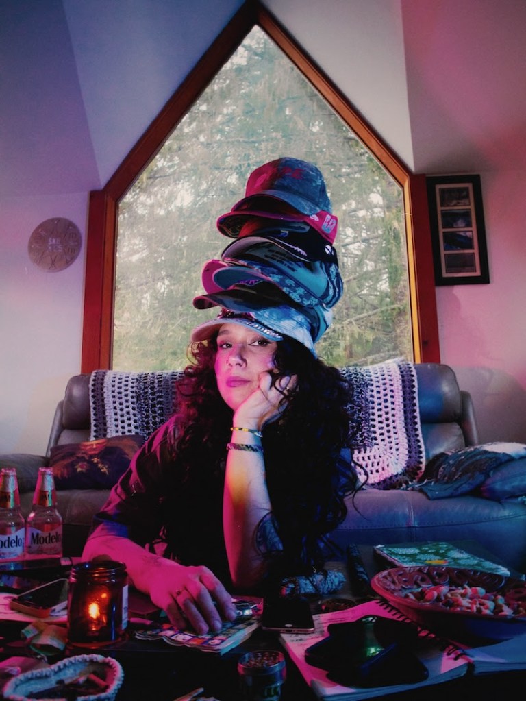 A young woman with curly hair sits at a table covered in various items, including snacks and drinks. She rests her chin on her hand, looking thoughtfully at the camera. On her head, a stack of colorful hats is balanced. The background features a large window with a view of trees, illuminated with soft colored lighting.