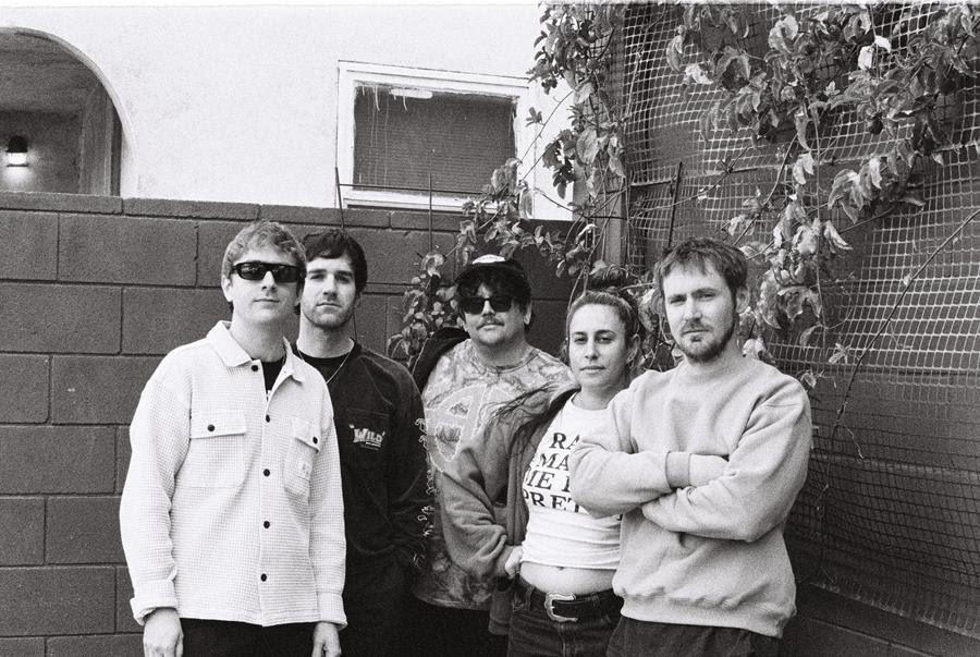 A group of five individuals posing together in a casual outdoor setting with greenery in the background, captured in black and white.